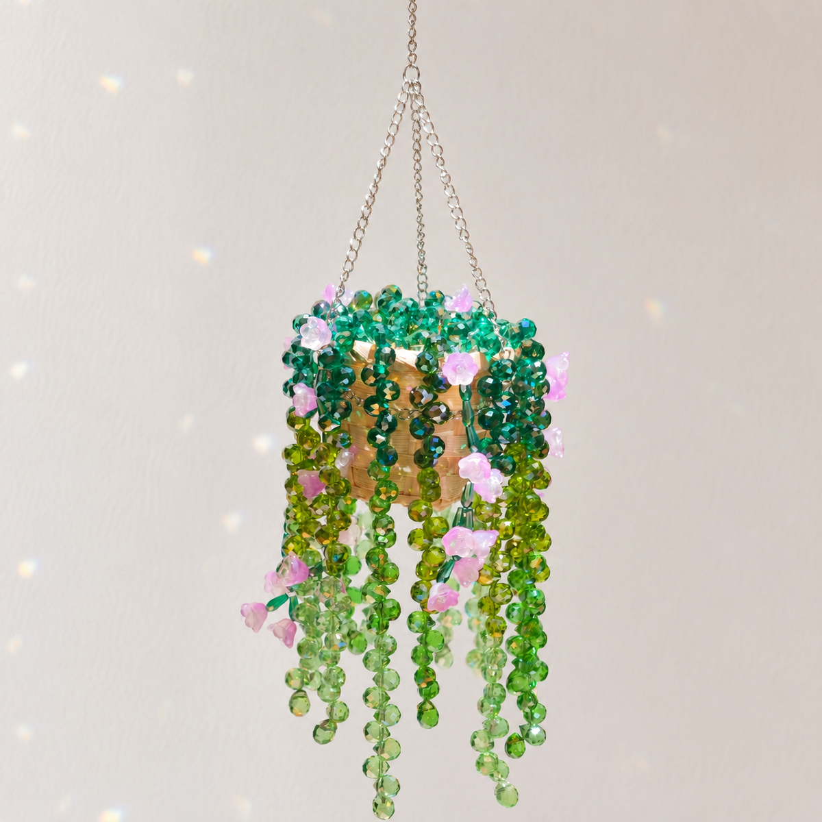 A hanging beaded plant ornament with green and light green faceted beads, pink flower accents, and a gold-toned center, suspended by chains against a light neutral background.