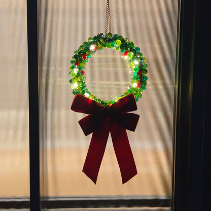 Green beaded Christmas wreath with warm fairy lights and a burgundy velvet bow hanging on a window