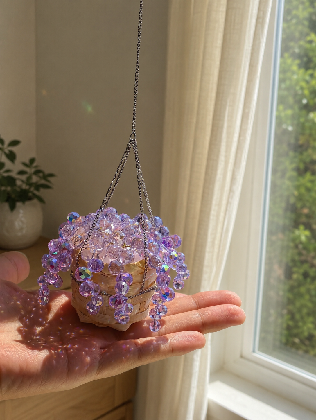 Hand holding a palm-sized finished purple beaded plant beside a window.
